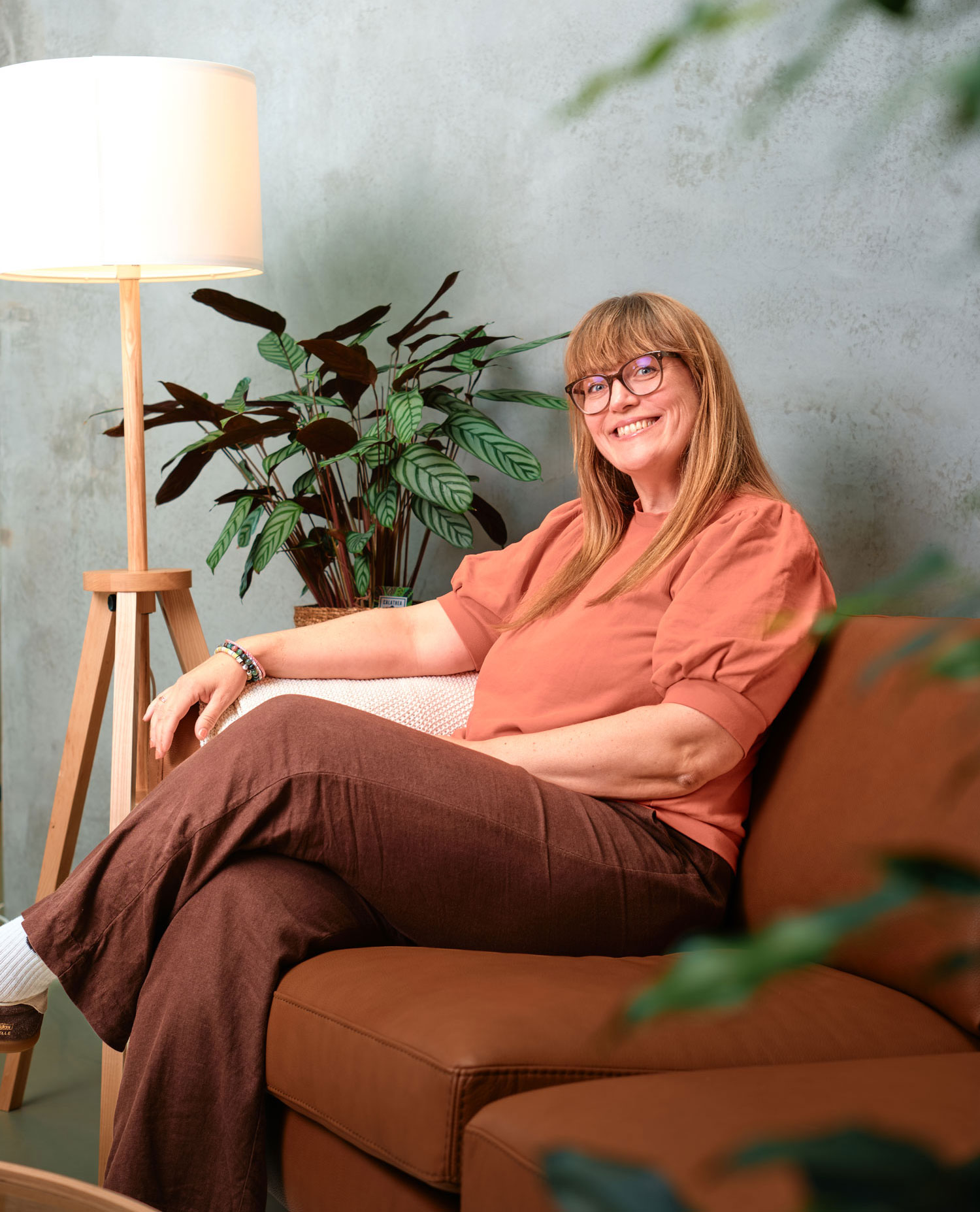 Aud Helen smiler i et profesjonelt portrettbilde, sittende i en brun sofa med en gulvlampe og en potteplante i bakgrunnen